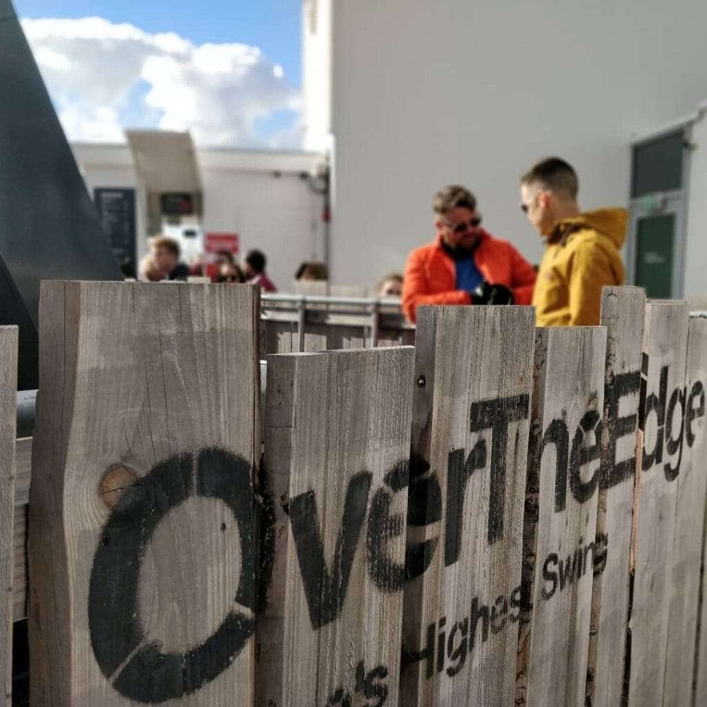 two men, one wearing an orange jacket and one wearing a yellow jacket standing behind a wooden fence with a stenciled sign saying Over The Edge.