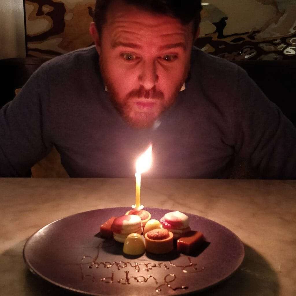 an image of a man blowing out a single candle on a small cake surrounded by other small cakes on a dark coloured plate