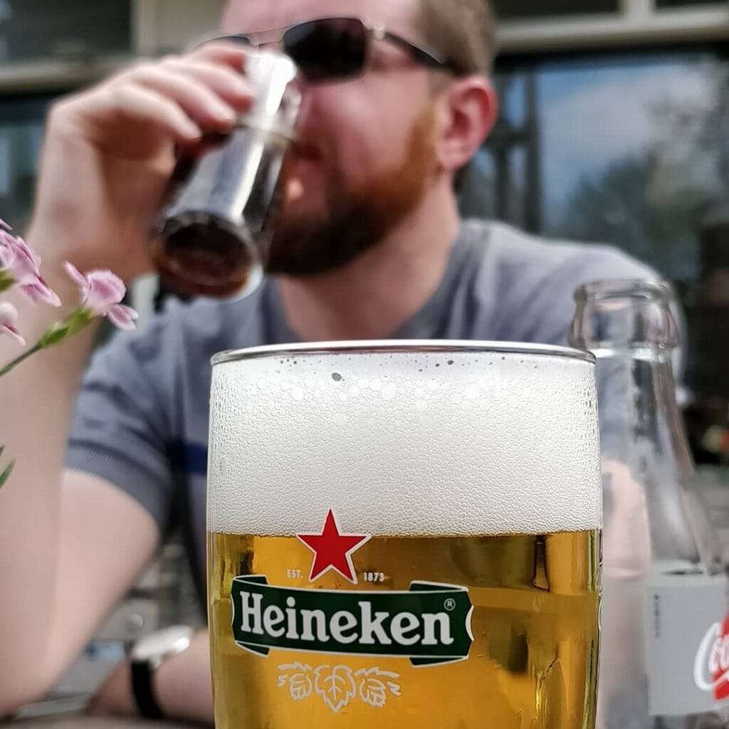 A portrait picture of a man drinking a glass of heineken he is our of focus and in the foreground in focus is a glass of heineken.