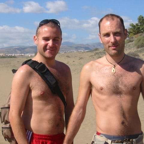 two topless men standing on sand dunes at maspalomas on the island of gran canaria. in the background are moutains and a blue but cloudy sky.