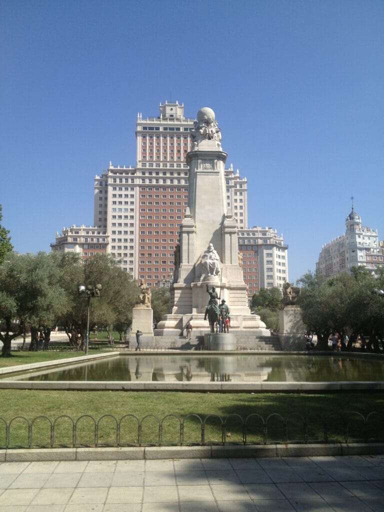 a photograph of the Monumento Cervantes in the foreground with the white and brick facade of Hotel Riu Plaza España behind it taken from Plaza de España in MAdrid with clear blue skies
