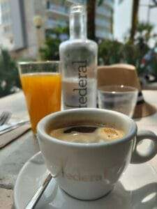a cup of coffee on a table with a glass of orange juice and a water bottle. in the background out of focus is a hotel and palm trees