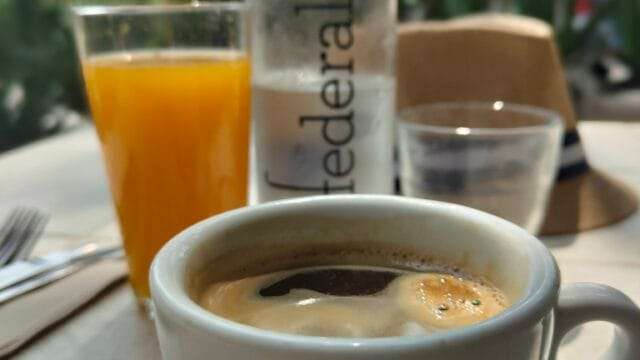 a cup of coffee on a table with a glass of orange juice and a water bottle. in the background out of focus is a hotel and palm trees